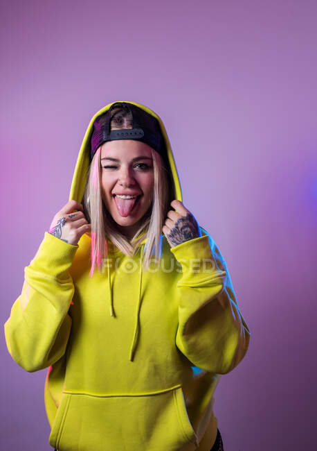 Rebelde hipster femenino en sudadera con capucha y gorra mirando a la cámara con la lengua en el fondo púrpura en el estudio con luces de neón - foto de stock