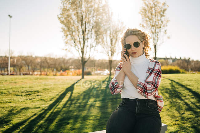 Mujer sonriente con gafas de sol sentada en un banco en el parque en un día soleado y hablando por teléfono móvil - foto de stock
