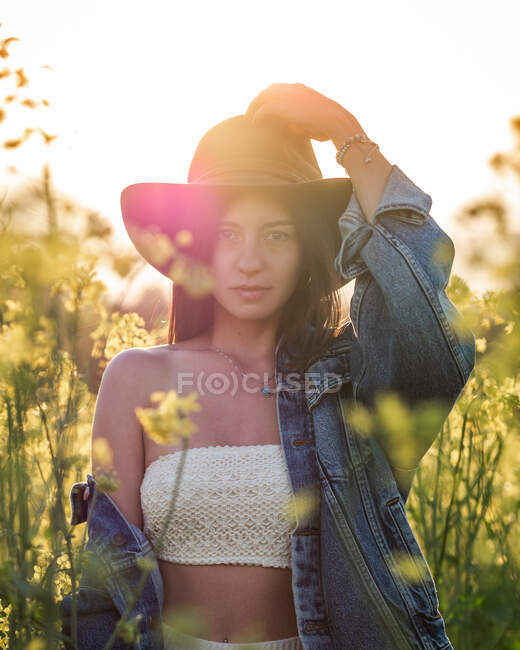 Young brunette in white top hat and lowered jacket standing looking at camera on blooming rapeseed field on sunny day — Stock Photo