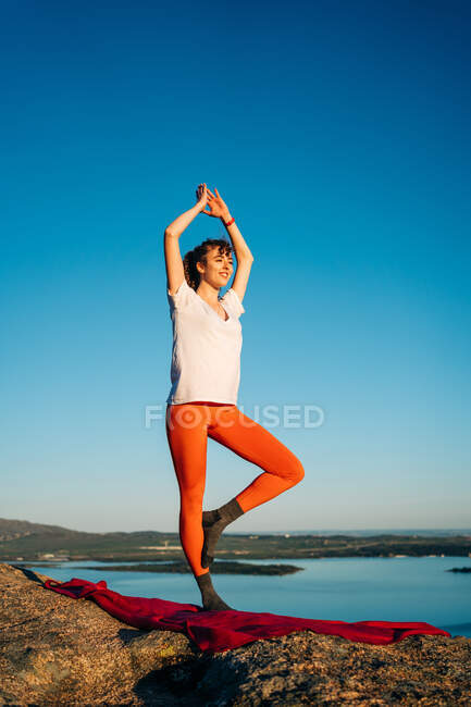 Young female traveler in sportswear standing in Tree pose with Arms Up asana while practicing yoga on rocky mountain over seacoast against cloudless blue sky — Stock Photo