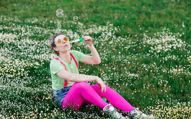 Виразна молода жінка в яскравому барвистому одязі грає з мильними бульбашками і сидить на зеленому лузі в природі — стокове фото
