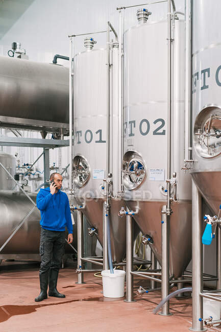 Vista lateral do empresário masculino falando no celular contra embarcações de aço inoxidável no chão molhado na fábrica de cerveja — Fotografia de Stock