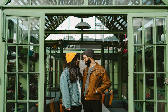 Cheerful couple of trendy hipsters standing on wooden terrace while holding hands and looking over shoulder — Stock Photo