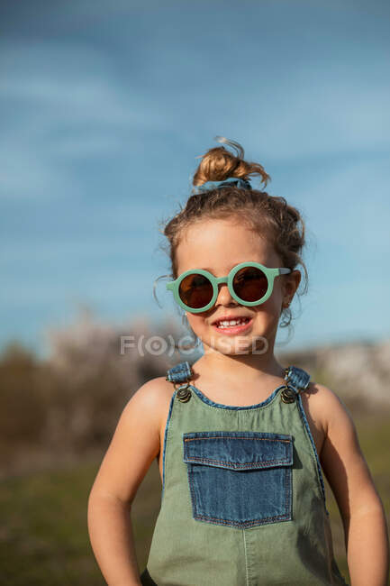 Zufriedenes kleines Mädchen in Overalls und Sonnenbrille steht auf der Wiese und genießt den Sommer an einem sonnigen Tag im Grünen — Stockfoto