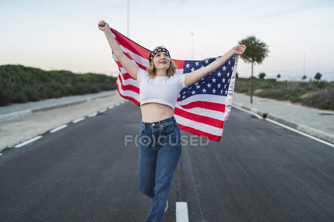 Donna americana deliziata in piedi con bandiera nazionale degli Stati Uniti sulla carreggiata al tramonto e guardando in alto — Foto stock
