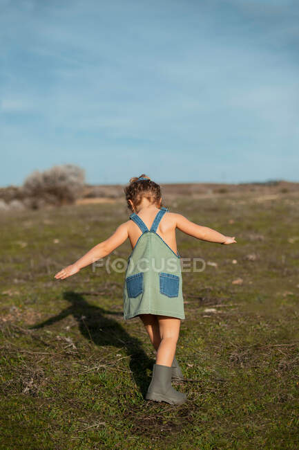 Back view adorable little girl in overalls standing with extended arms in meadow and looking down — Stock Photo