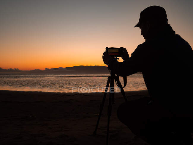 Side view of unrecognizable male photographer standing near tripod with photo camera and preparing for shooting seascape at sundown time — Photo de stock