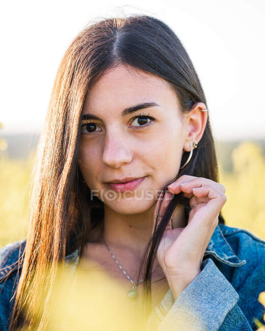 Young brunette in white top and denim jacket standing looking at camera on blooming rapeseed field on sunny day — Stock Photo