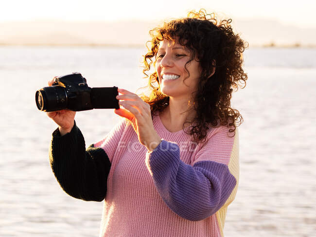Весела молода кучерява етнічна жінка мандрівник перевіряє фотографії на фотоапараті, стоячи біля моря в сонячний вечір — стокове фото