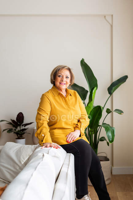 Задоволена зріла жінка сидить на дивані і дивиться на камеру в затишній вітальні з кімнатними рослинами — стокове фото