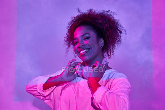 Fröhliches Porträt einer jungen Afroamerikanerin in weißer Kleidung, die Kopfhörer am Hals trägt und in die Kamera schaut, während sie im dunklen Studio im Neonlicht steht — Stockfoto