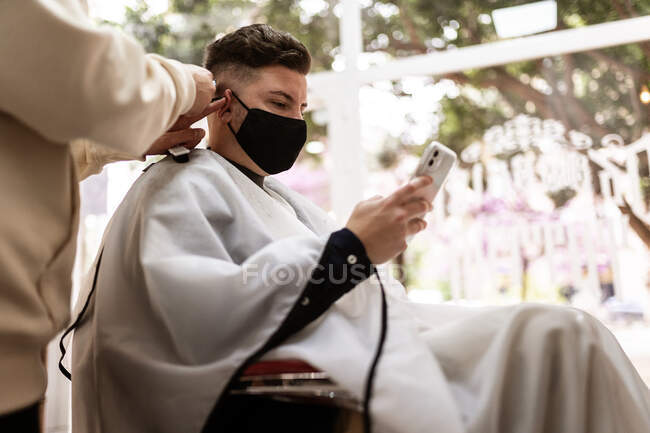 Снизу стилист урожая с триммером стрижка волос человека в тканевой маске лица с сотовым телефоном в парикмахерской — стоковое фото