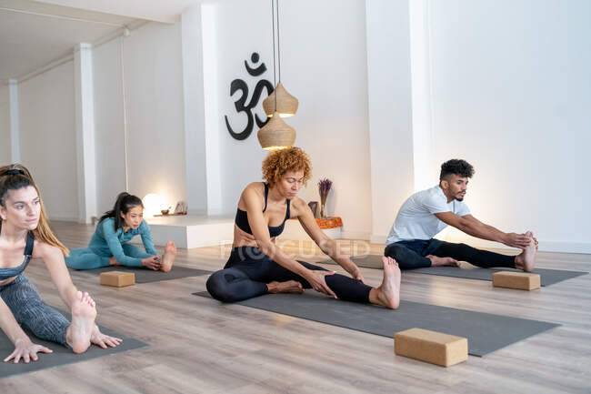 Empresa de personas multiétnicas enfocadas en ropa deportiva sentadas sobre esteras en pose de flexión hacia adelante y practicando yoga juntas en estudio - foto de stock