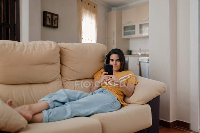 Young female text messaging on cellphone while lying down on couch in living room — Stock Photo