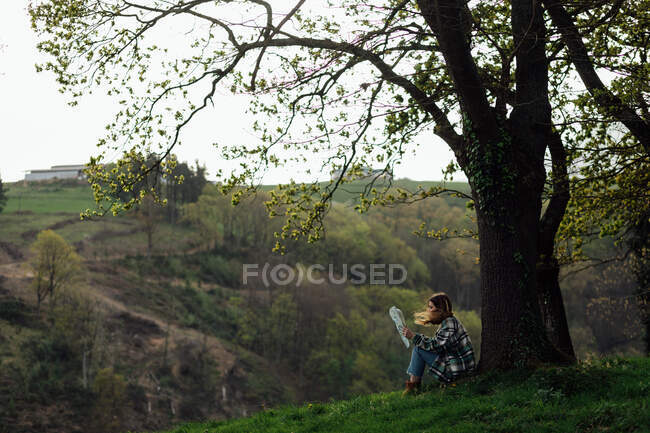 Side view of female traveler with paper guide sitting on meadow between green trees and mount in countryside — Photo de stock