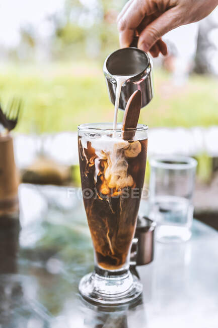 Cultiver barista méconnaissable verser du lait frais du pichet dans un verre de café noir froid servi sur une table en verre dans la cafétéria extérieure — Photo de stock