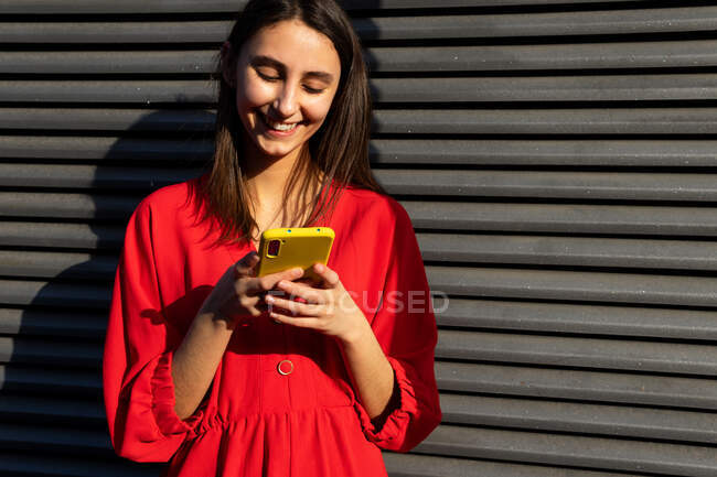 Joven contenido femenino en rojo desgaste chat en el teléfono móvil en la luz del sol sobre fondo gris - foto de stock