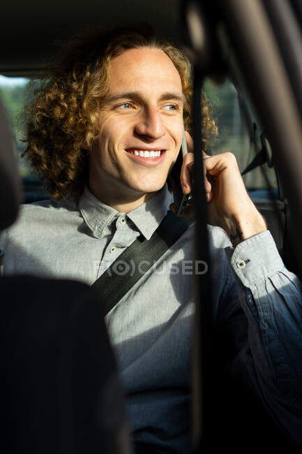Cheerful young stylish haired male sitting on driver seat and speaking on mobile phone during trip through nature in sunny summer day — Fotografia de Stock