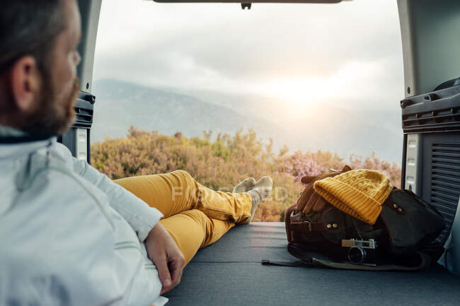 Vista trasera de un campista masculino irreconocible en ropa de abrigo sentado en la furgoneta y admirando la vista panorámica de las tierras altas - foto de stock