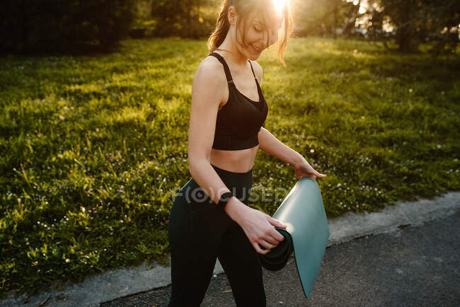 Vista laterale di atleta femminile in forma di raccolto in piedi con stuoia nel parco estivo mentre si prepara per l'allenamento in serata — Foto stock