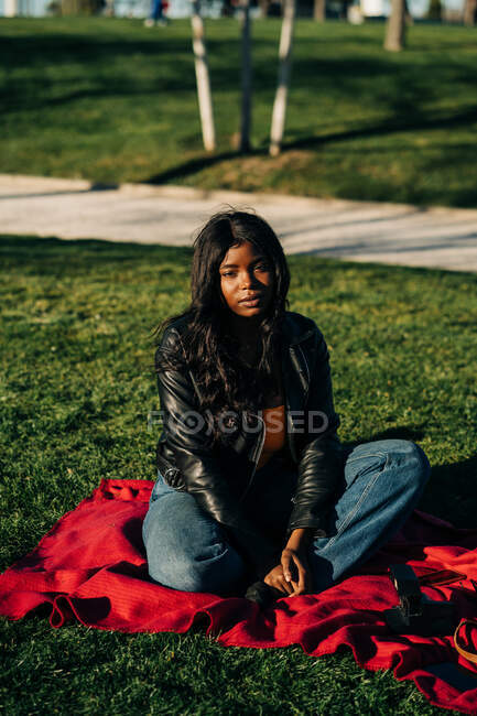 Unbekümmerte Afroamerikanerin im schicken Outfit sitzt auf kariertem Rasen im Park und blickt in die Kamera — Stockfoto