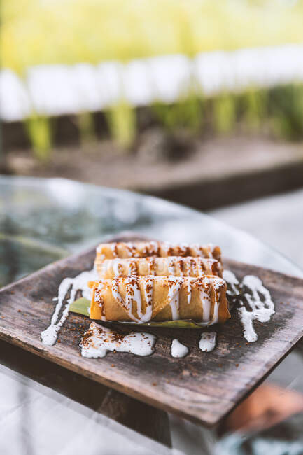 Deliziosi involtini dolci con salsa alla vaniglia serviti su un piatto di legno su un tavolo di vetro nel ristorante asiatico all'aperto — Foto stock