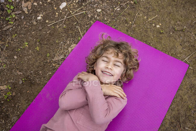 From above of content child with crossed arms and wavy hair lying on bright mat in daytime outdoors - foto de stock