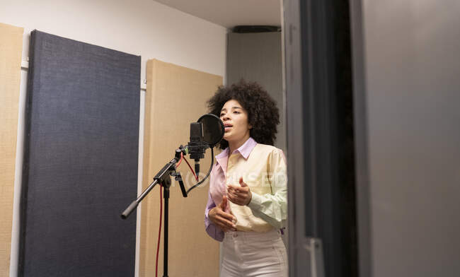 Cantante femenina negra interpretando canción contra micrófono con filtro pop mientras está de pie con la mano en la cadera y mirando hacia adelante en el estudio de sonido - foto de stock