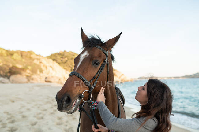 Young female walking and caressing chestnut stallion in bridle against wavy ocean — Stock Photo