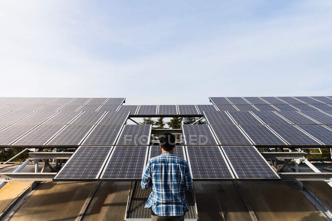 Volver ver anónimo técnico profesional con camisa a cuadros comprobación de paneles fotovoltaicos en la central solar en tiempo claro y soleado - foto de stock
