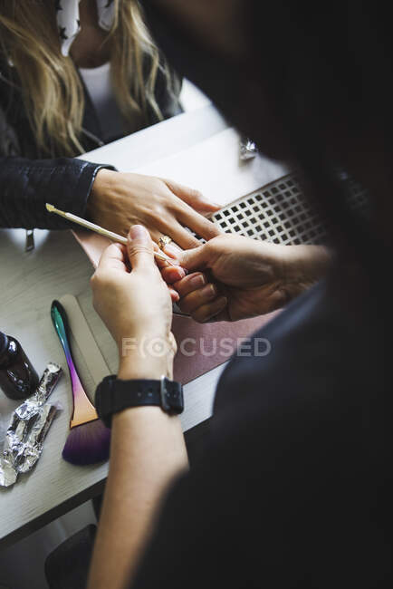 Alto ángulo de cultivo anónimo belleza maestro de uñas de la mujer que utiliza el tablero de esmeril durante el procedimiento de manicura en la mesa - foto de stock