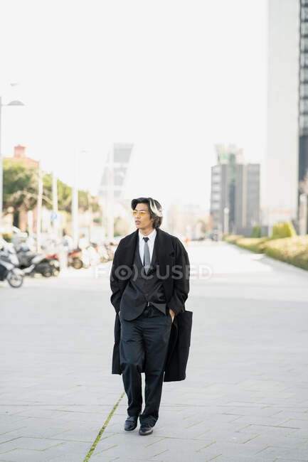 Empresario masculino étnico joven y seguro de sí mismo con traje y abrigo caminando con las manos en los bolsillos mientras mira hacia otro lado en la ciudad - foto de stock
