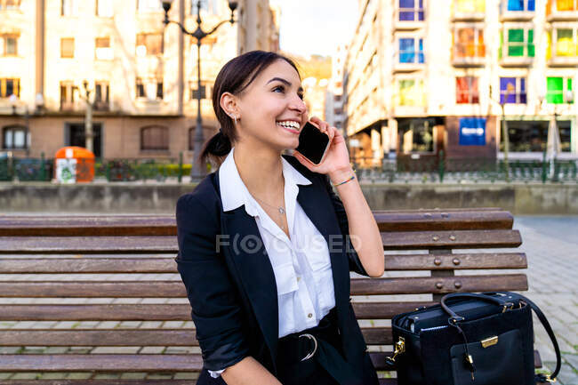 Zufriedene junge ethnische Unternehmerin sitzt auf Bank, während sie auf dem Smartphone spricht und wegschaut von Elektroroller und städtischen Gebäuden — Stockfoto