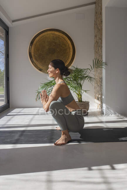 Vista lateral de la joven hembra en ropa deportiva mostrando la pose de Malasana mientras practica yoga con los ojos cerrados en casa - foto de stock