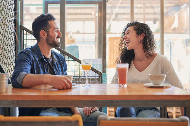 Alegre pareja étnica mirándose mientras se divierten en la mesa de madera con café y bebidas refrescantes - foto de stock