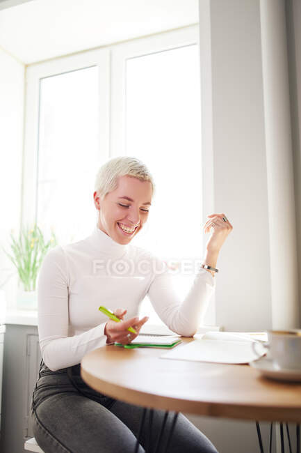 Vista lateral da astróloga feliz tomando notas no bloco de notas na mesa com xícara de café em casa à luz do sol — Fotografia de Stock