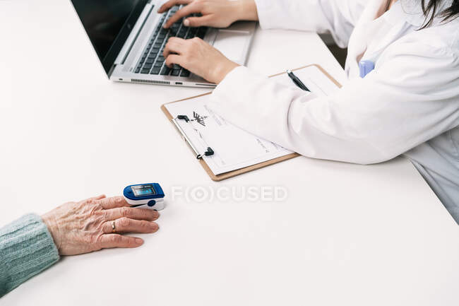 Crop unrecognizable doctor measuring blood oxygen of patient with heart rate monitor at table with stethoscope in clinic while using laptop — Stock Photo