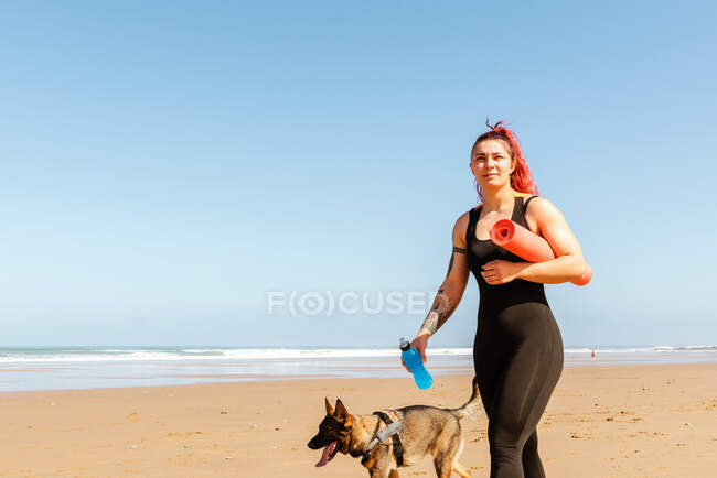 Lächelnde Athletin mit Rollmatte und Wasserflasche spaziert mit reinrassigem Hund an der Sandküste und schaut weg — Stockfoto
