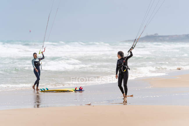 Multiethnic female athletes in wetsuits with kiteboard and control bars looking at each other on sandy coast against foamy ocean — Stock Photo