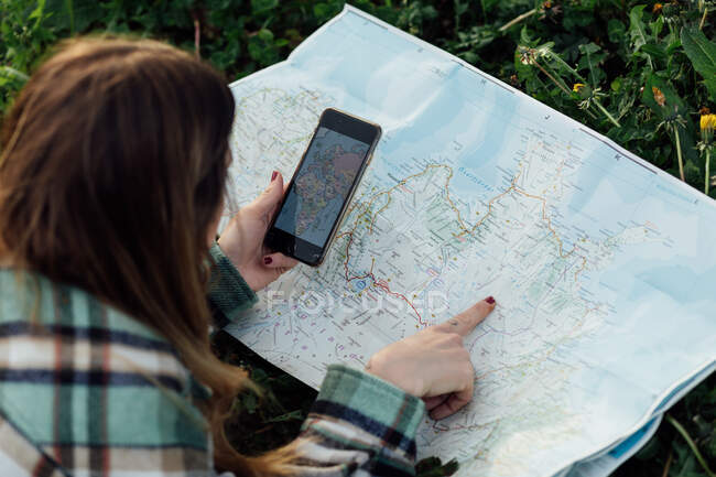 Сверху сзади вид неузнаваемой туристки с картой маршрута наблюдения за мобильным телефоном, лежащей на траве в сельской местности — стоковое фото