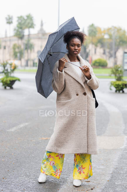 Cuerpo completo joven mujer afroamericana de moda en abrigo cálido de pie con paraguas en la calle de la ciudad moderna y mirando a la cámara - foto de stock