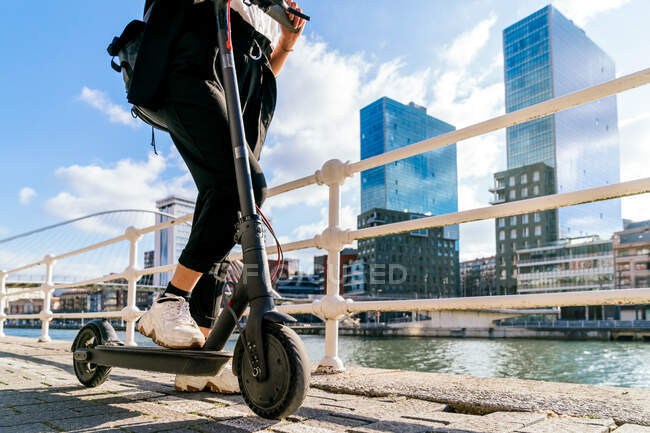 Side view of crop anonymous female in sneakers riding modern scooter on footpath against city river — Stock Photo