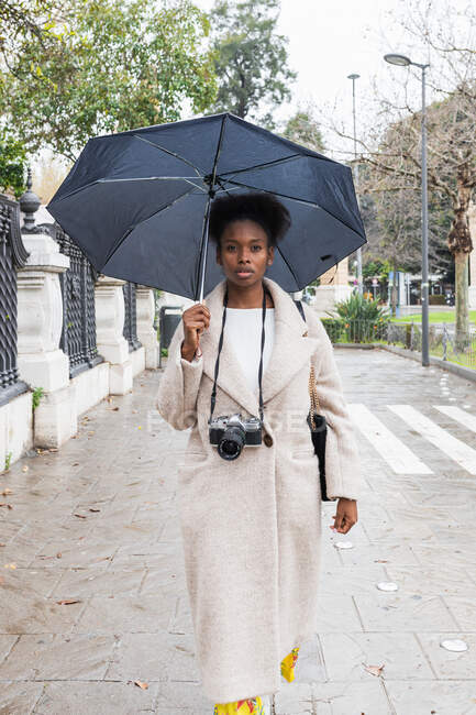 Joven mujer afroamericana de moda con abrigo cálido de pie con paraguas en la calle de la ciudad moderna y mirando a la cámara mientras usa una cámara profesional - foto de stock