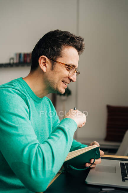 Vista lateral do trabalhador remoto masculino alegre tomando notas contra netbook com tela preta enquanto trabalhava na mesa em casa — Fotografia de Stock