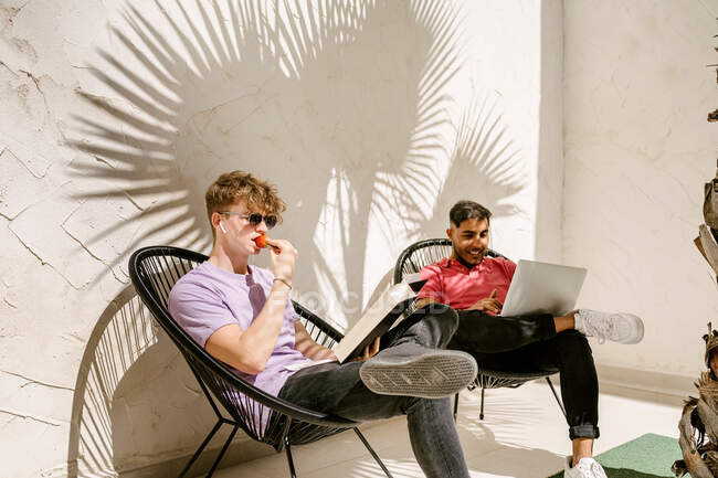 Стильні молоді багаторасові друзі-чоловіки використовують ноутбук і читають книгу, відпочиваючи на стільцях на терасі в сонячний день — стокове фото