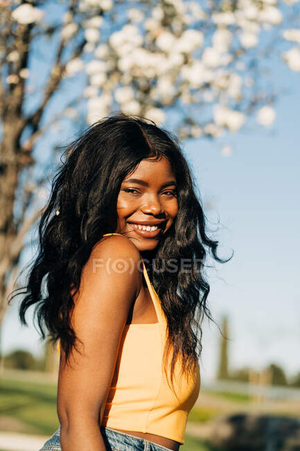 Beautiful cheerful African American female standing in blooming spring park and enjoying sunny weather looking at camera — Stock Photo