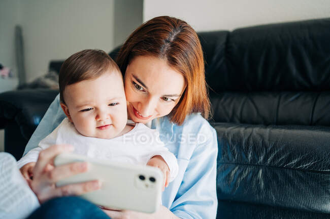Jovem mulher tomando selfie com a criança encantadora no telefone celular enquanto passa o tempo em casa — Fotografia de Stock