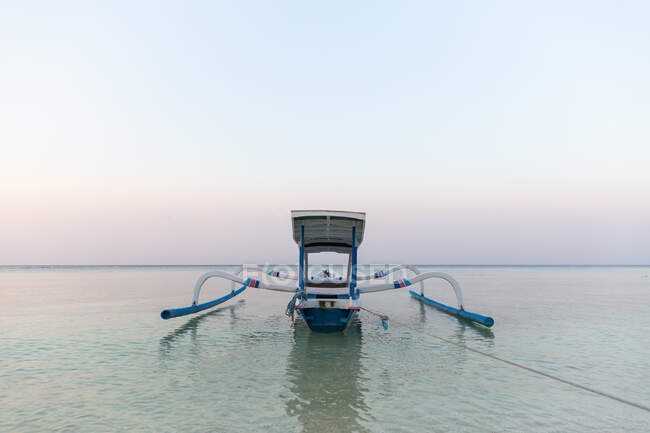 Маленькая рыбацкая лодка пришвартована на бирюзовой морской воде под безоблачным голубым небом в мирных сумерках — стоковое фото