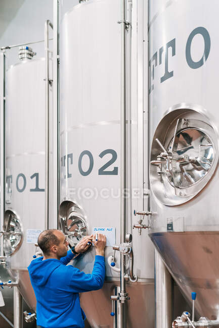 Задний вид мужчины-инженера, делающего заметки на бумаге на металлическом судне на пивоваренном заводе — стоковое фото