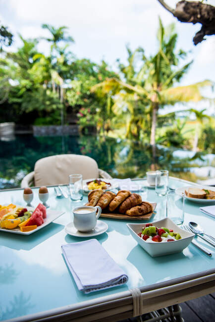 From above of comfortable wicker armchair with soft seats and cushions placed near table served with various appetizing pastries and fruits during breakfast in tropical resort — Photo de stock